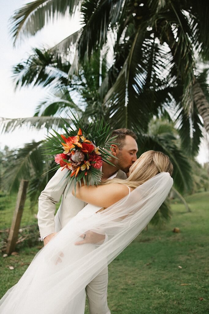 A bride is dipped and kissed by the groom while standing in front of palm trees