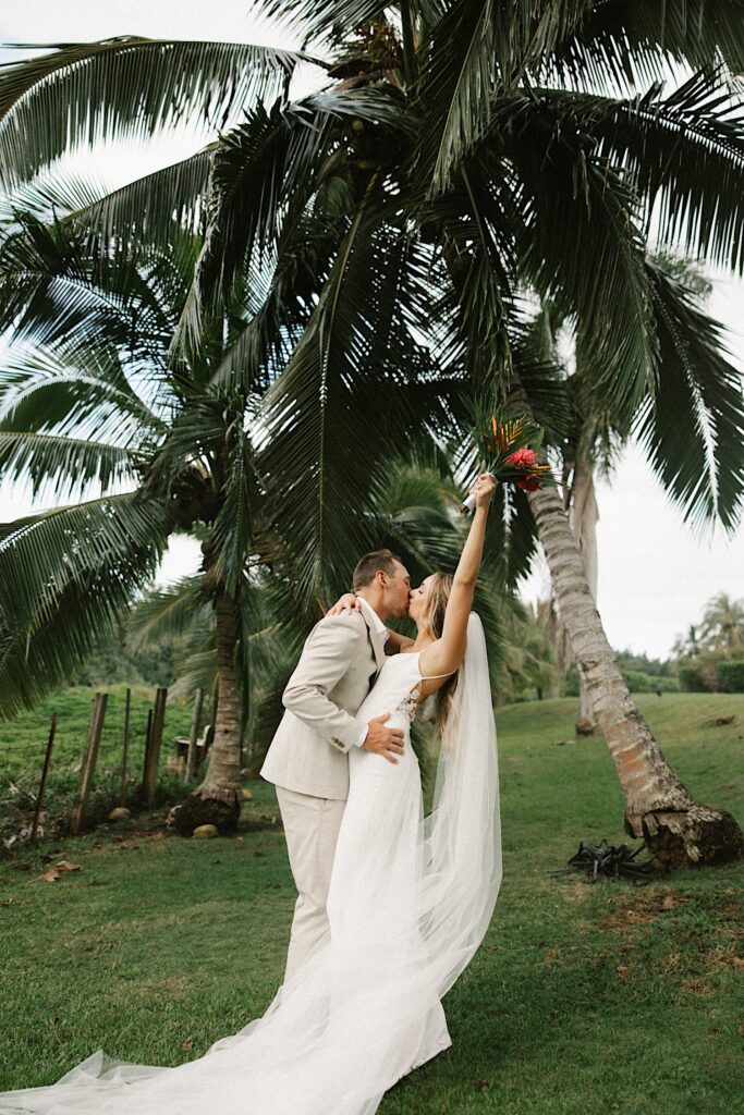 A bride and groom kiss in front of palm trees while the bride raises her bouquet in the air