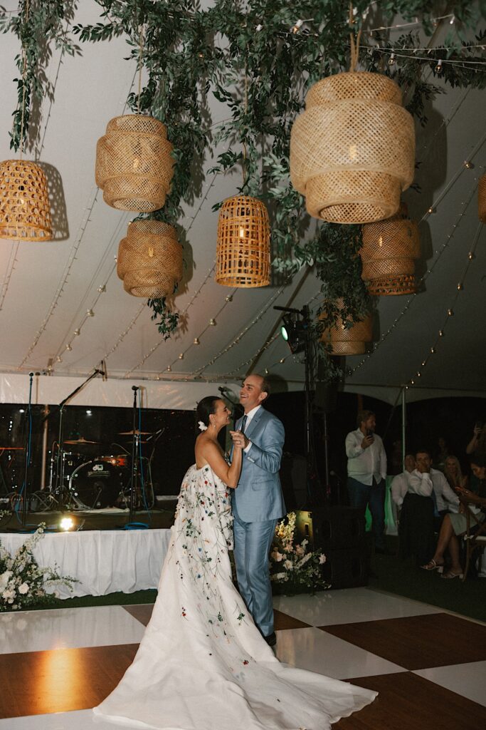 A bride and groom share their first dance underneath a tent decorated with string lights, greenery, and lanterns