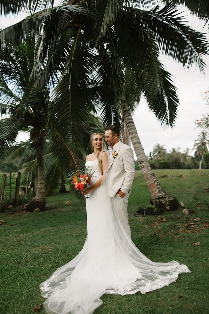 A bride stands in front of a groom and looks back at him over her shoulder while the two stand in front of palm trees