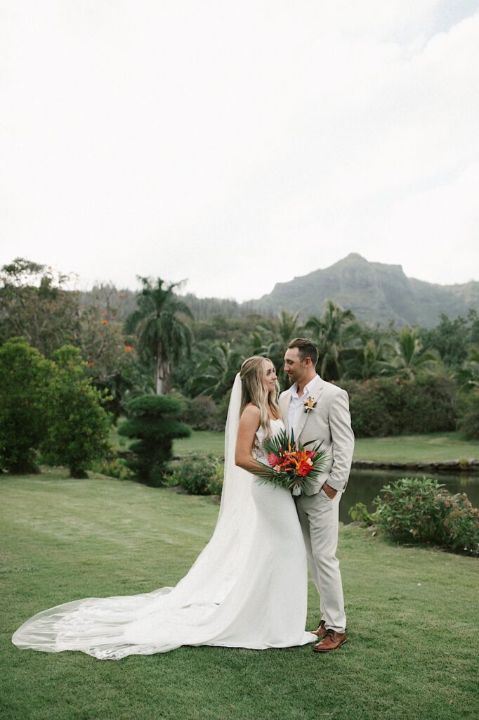 A bride and groom embrace and look at one another while standing in front of a lake, palm trees, and a mountain