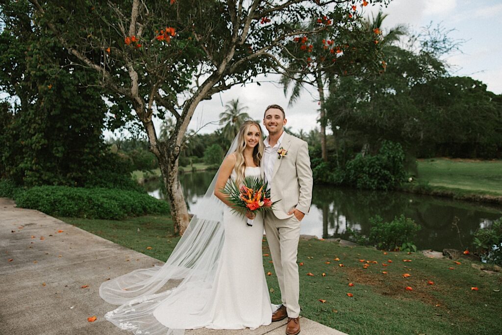 After their intimate wedding ceremony on Kauai, a bride and groom stand next to one another and smile at the camera with a small pond and trees behind them