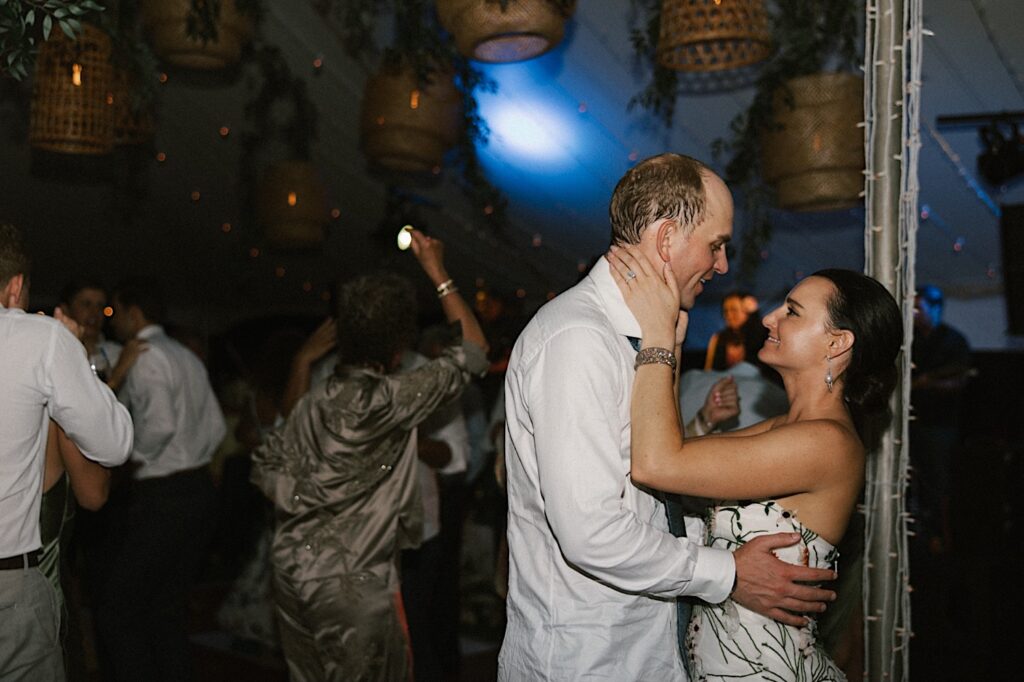 A bride and groom dance underneath a tent decorated with string lights with the guests of their wedding reception around them at their wedding venue Kukui'ula on Kauai