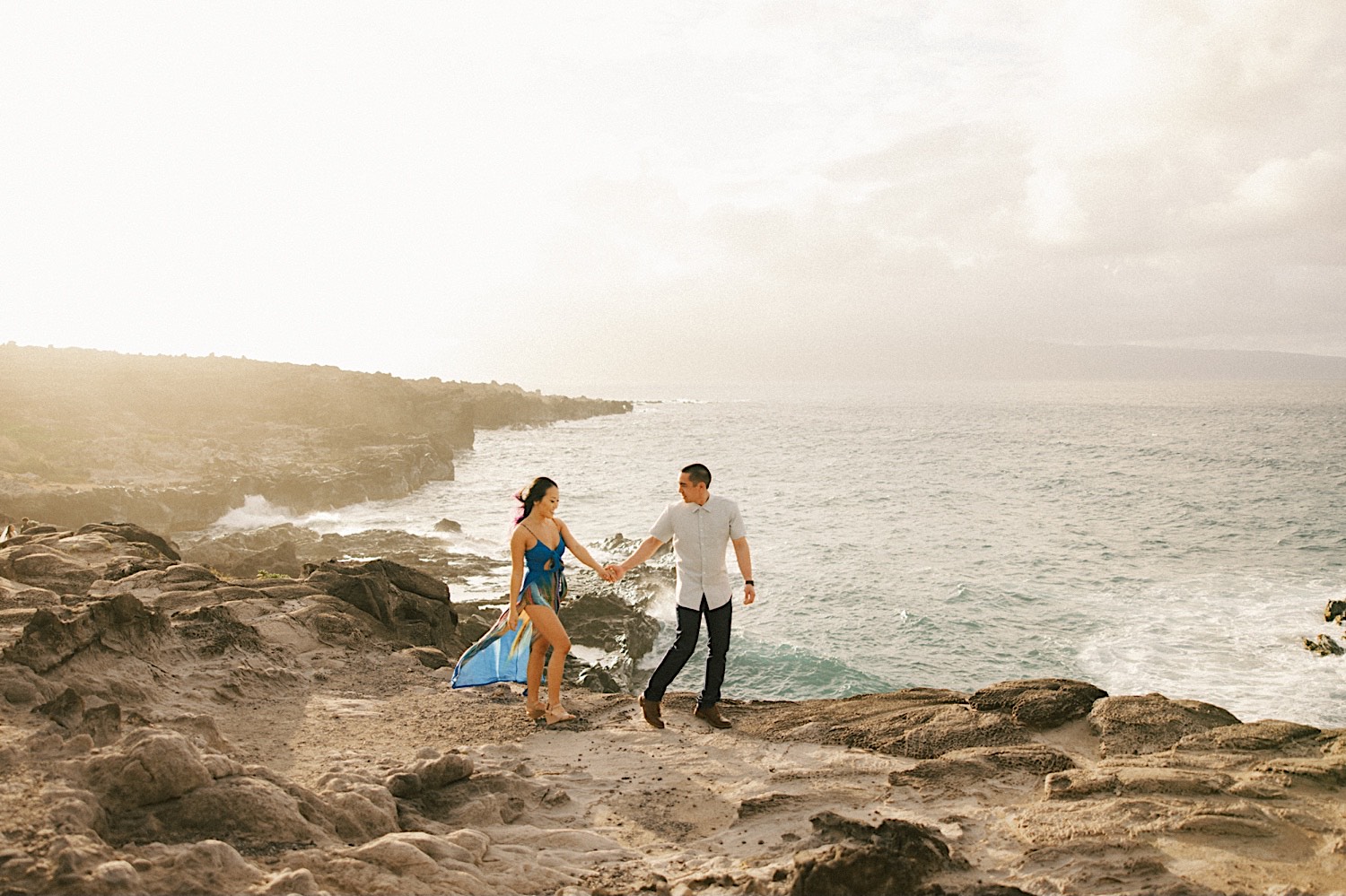 During their engagement session on the island of Maui a man and woman walk hand in hand on a cliff looking out over the ocean below them as the sun sets to the left
