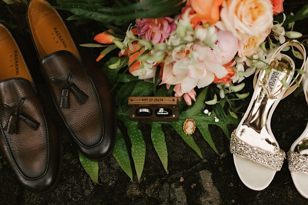 Two wedding rings sit in a wooden box with a wedding date on them, on either side of the box are a man's and woman's shoes, also on the ground are leaves, flowers, and earrings
