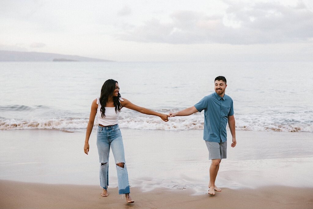 During their engagement session on the island of Maui a man and woman hold hands and smile while walking on the beach towards the camera with the ocean behind them
