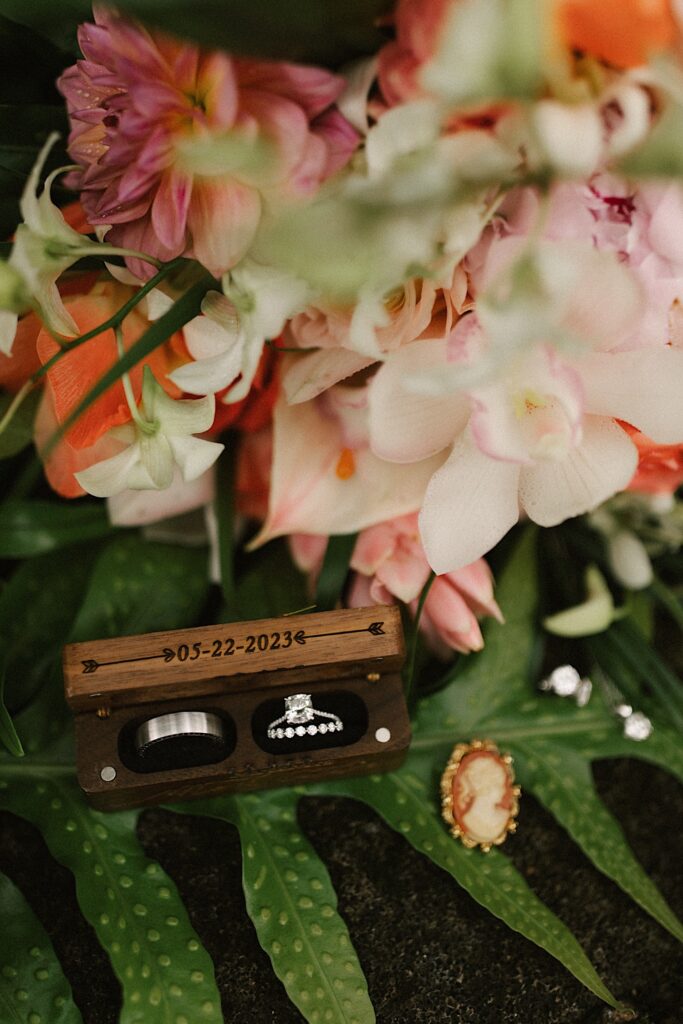 2 wedding rings in a wooden box with the date "05-22-2023" lay on a leaf in front of florals and with earrings next to it