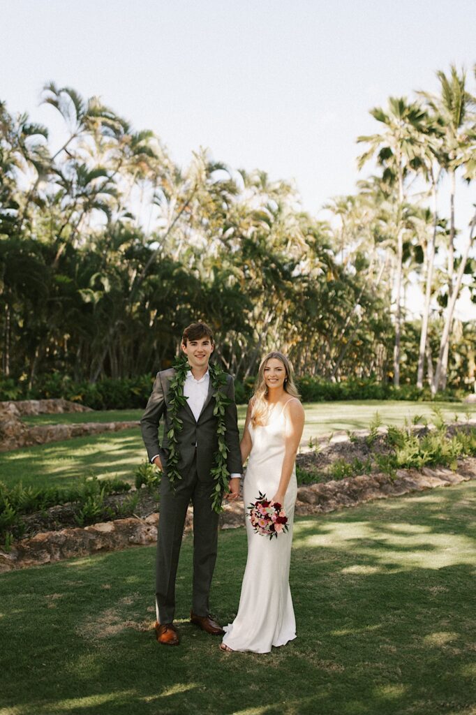 A bride and groom stand next to one another and smile at the camera with palm trees in the background before their elopement ceremony
