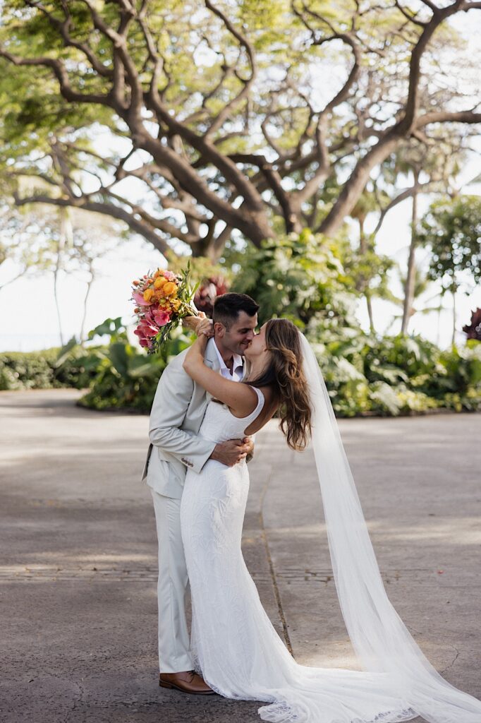 A bride and groom kiss one another while standing on a road underneath a giant tree