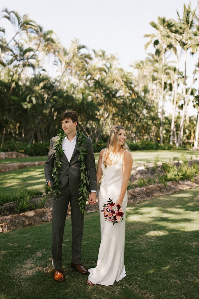 A bride and groom stand next to one another and hold hands while looking in opposite directions with palm trees behind them before their elopement ceremony