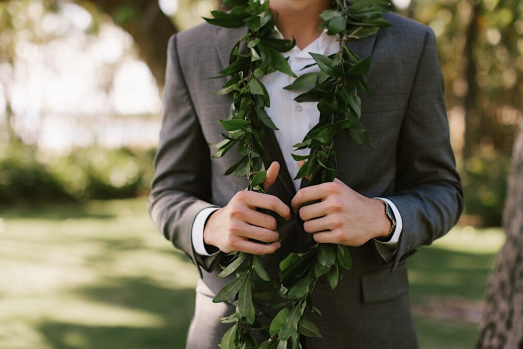 Shoulder down photo of a groom with a leaf lei around his next standing in front of trees