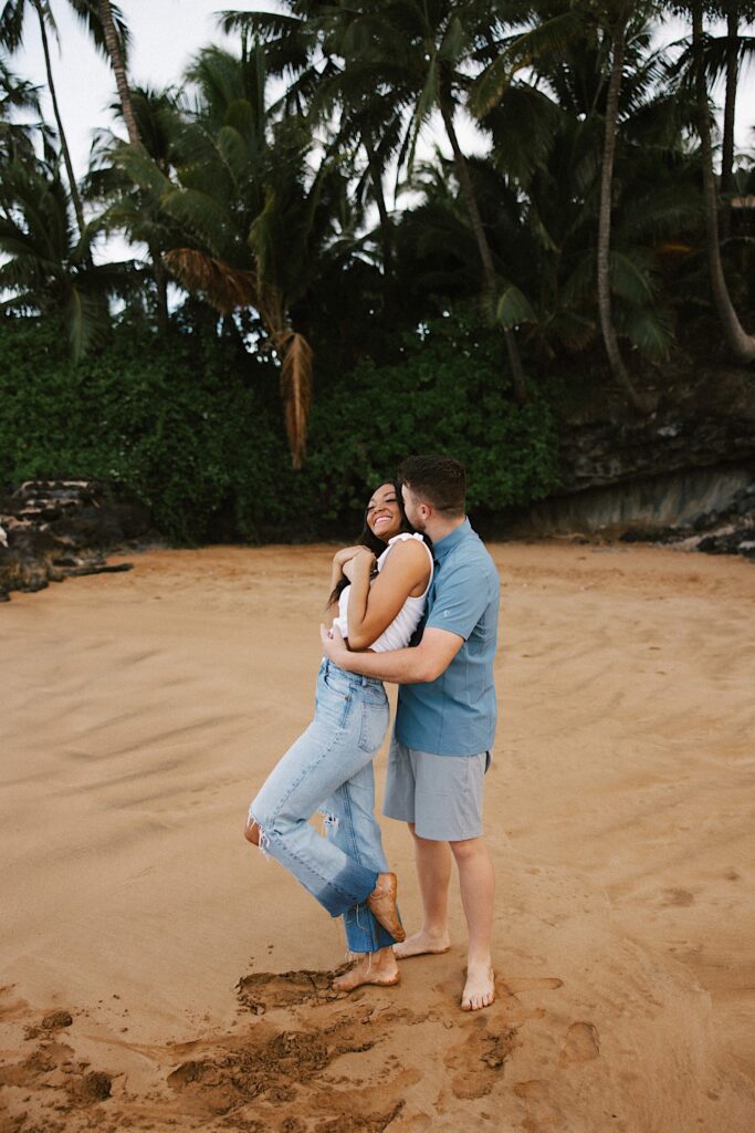 A woman smiles as a man hugs her from behind and kisses her on the cheek while they stand on a beach with palm trees behind them