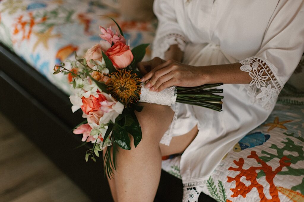A floral bouquet sits on the lap of a bride sitting on a bed before her wedding day
