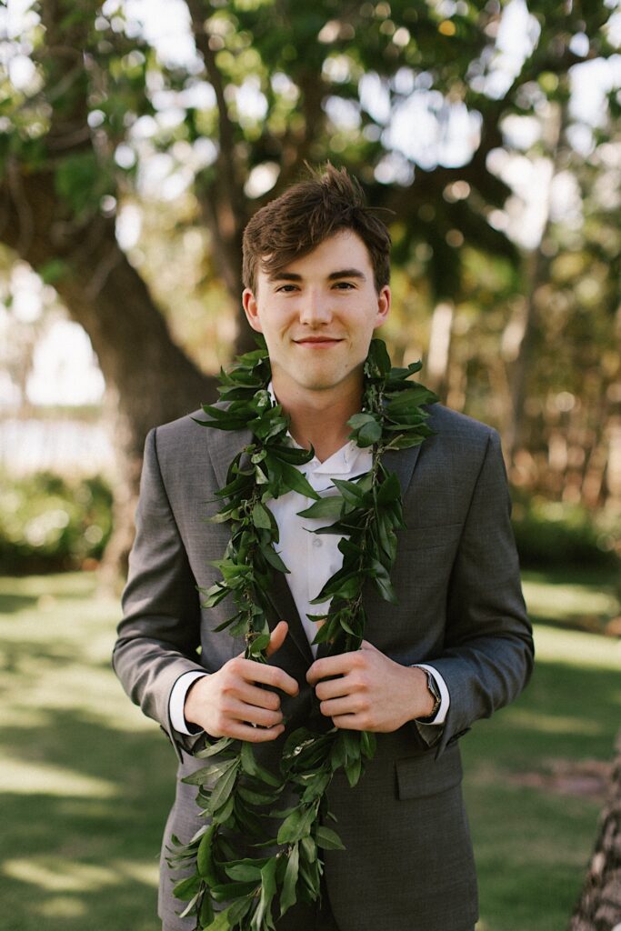 Portrait photo of a groom with a leaf lei around his neck looking at the camera with other trees behind him
