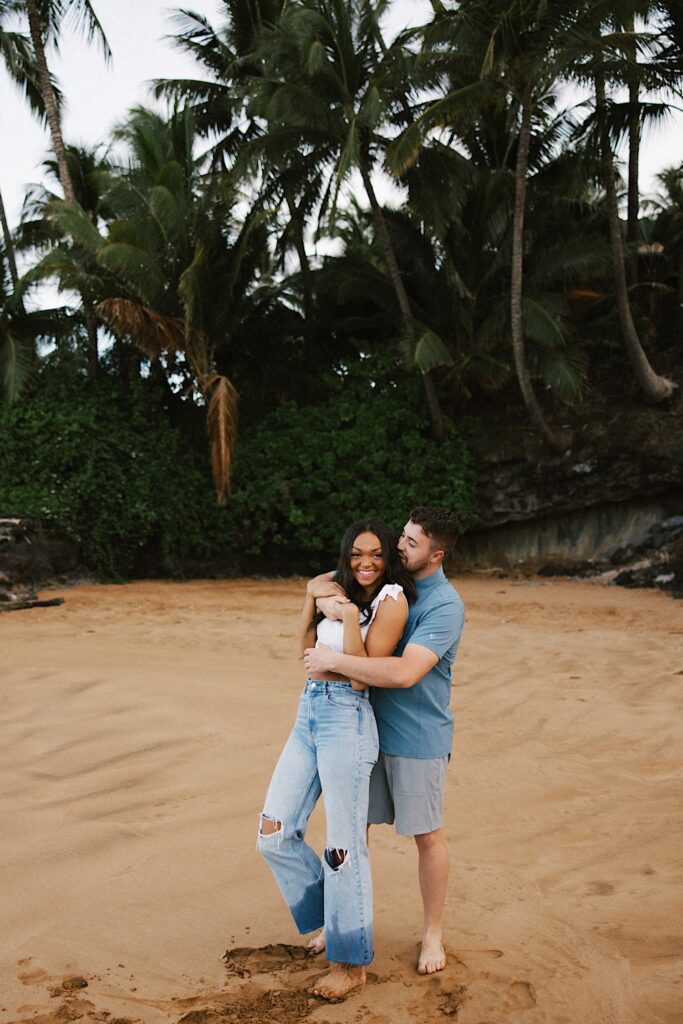 A woman smiles as a man hugs her from behind while they stand on a beach with palm trees behind them