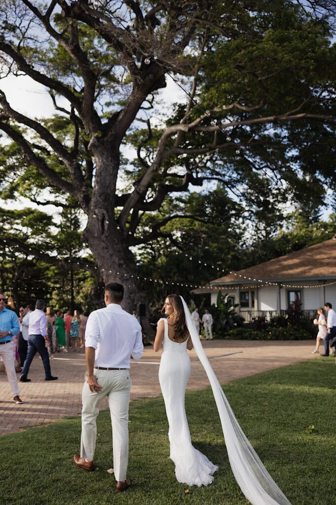 A bride and groom walk side by side away from the camera towards their wedding reception taking place under a massive tree
