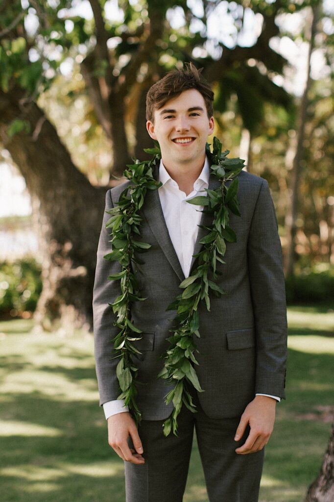 Portrait photo of a groom standing and smiling at the camera in front of trees with a leaf lei around his neck