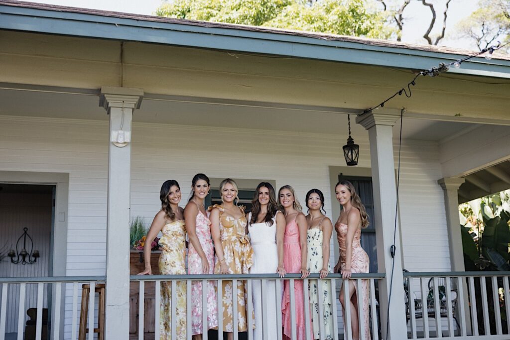 A bride stands with her 6 bridesmaids on either side of her on a front porch of a house as they all smile at the camera