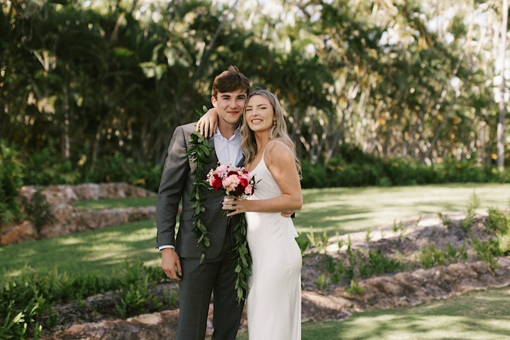 A bride and groom smile at the camera while embracing one another during their elopement at Lanikūhonua on Oahu, behind them are palm trees and a field of grass