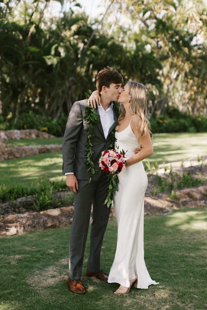 A bride and groom kiss one another while standing beside each other, behind them are palm trees and a field of grass