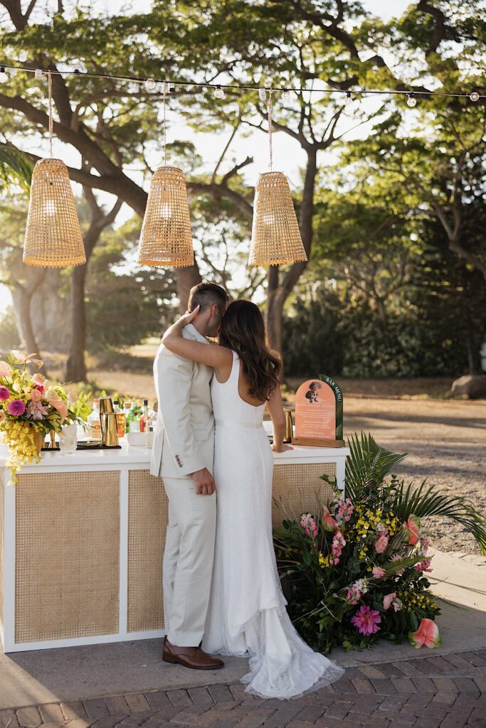 A bride and groom embrace while standing next to one another facing away from the camera looking at their drink table with trees and lush greenery all around them