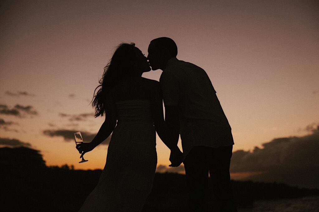 Silhouette of a couple kissing with the sun setting behind them and the woman is holding a glass of champagne