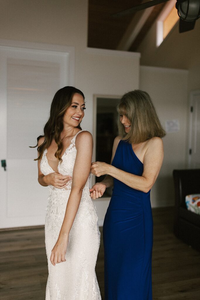A bride in her wedding dress smiles over her shoulder as her mother helps zip up the back of her dress