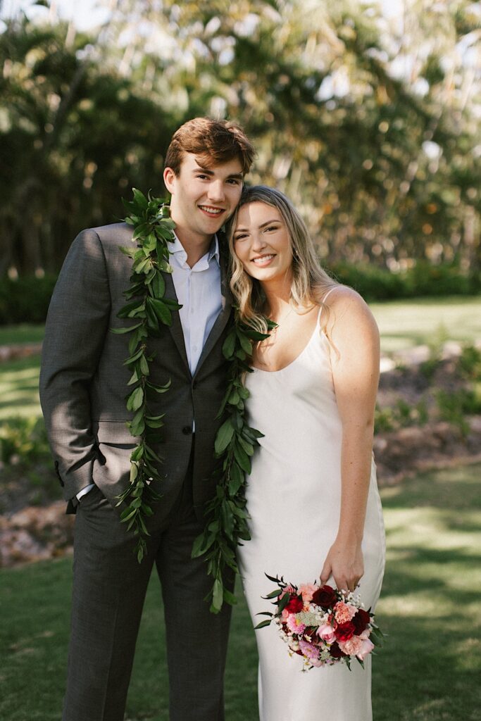 A bride and groom stand side by side and smile at the camera, the bride is holding a bouquet and the groom is wearing a leaf lei, behind them are palm trees