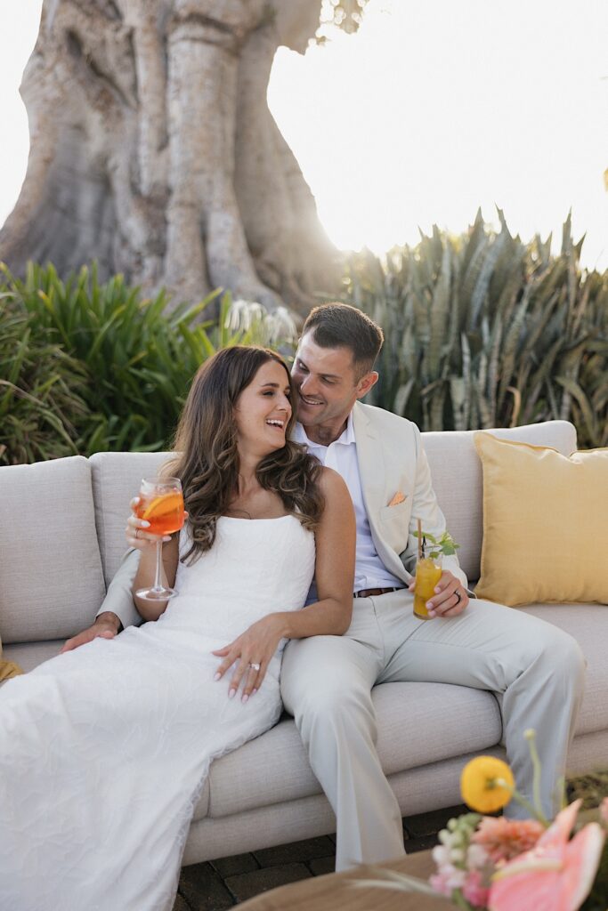 A bride and groom sit on a couch and each have a drink in hand while the bride leans into the groom and they both smile at one another with plants and a giant tree behind them