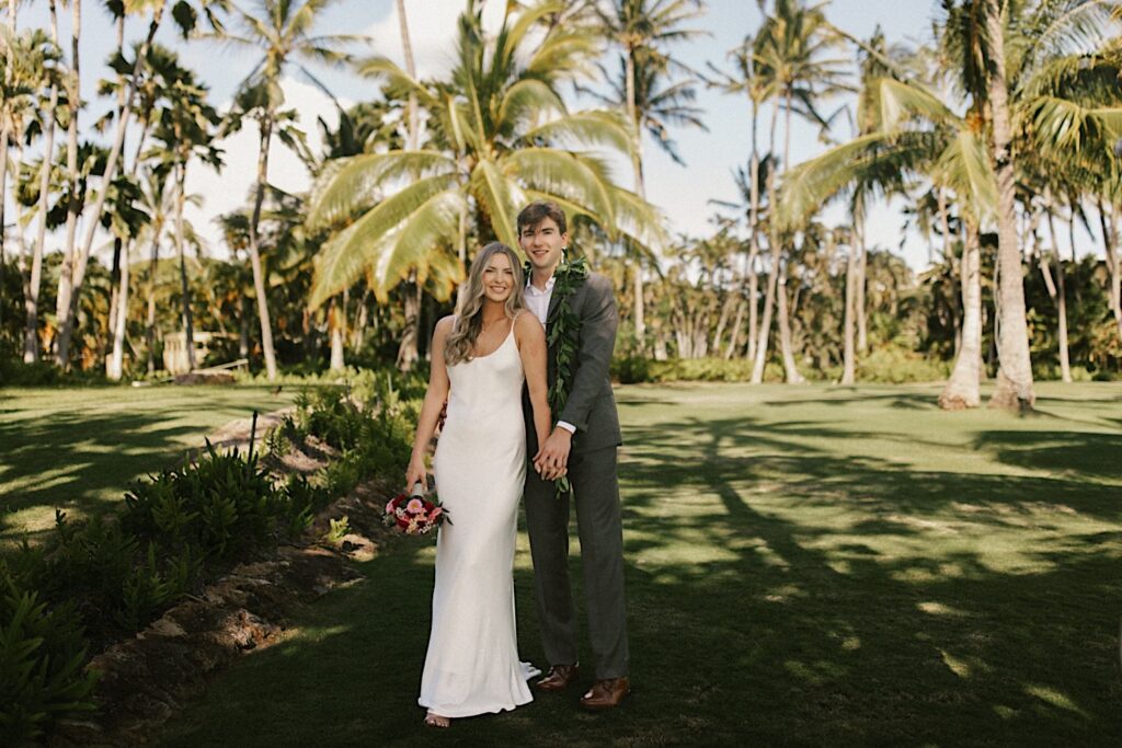 A bride and groom stand together during their elopement at Lanikūhonua on Oahu and hold hands while smiling at the camera, behind them are palm trees