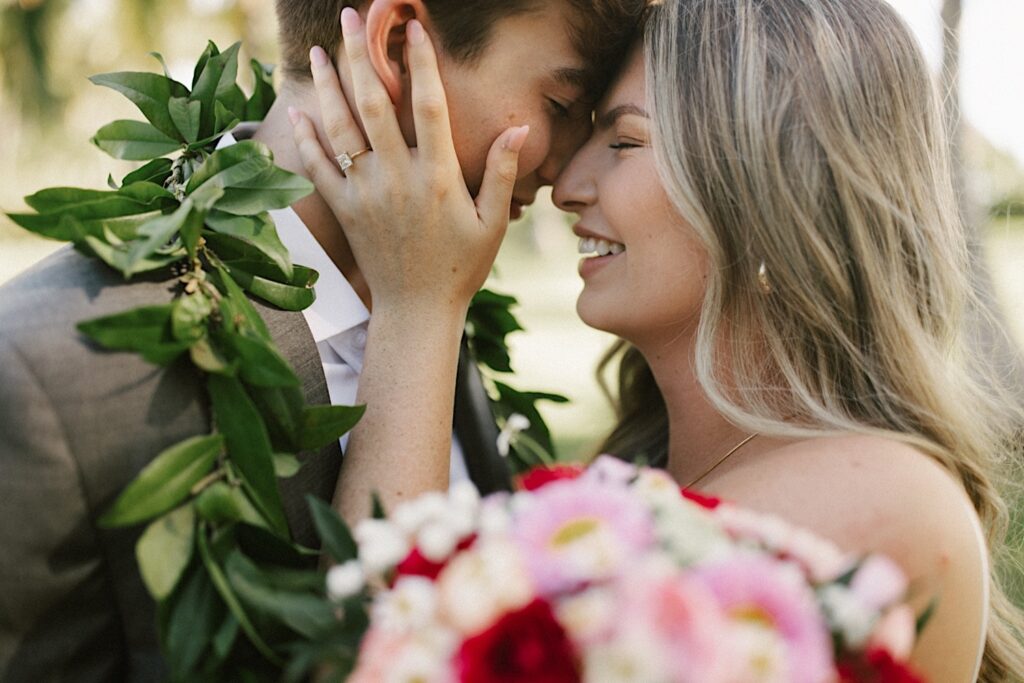 Close up photo of a bride smiling while holding the groom's face as the two touch their foreheads together during their elopement at Lanikūhonua on Oahu