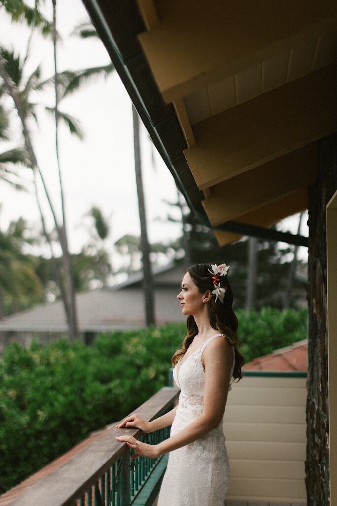 A bride standing on an outdoor patio leans on a railing looking out over the ocean with palm trees behind her
