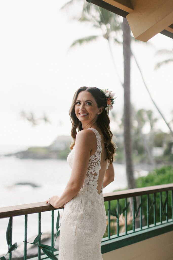 A bride leans on a railing looking out over the ocean and smiles back over her shoulder to the camera behind her