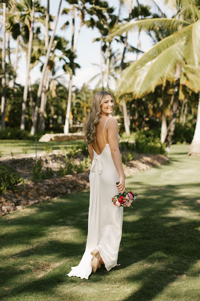Portrait photo of a bride with her back to the camera smiling over her shoulder  while holding her bouquet, behind her are palm trees