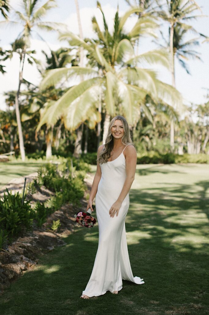 Portrait photo of a bride facing the camera and smiling while holding her bouquet, behind her are palm trees