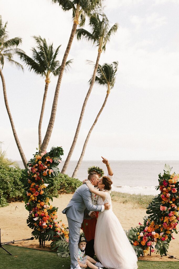 A bride and groom kiss one another while their officiant cheers behind them, also behind them are palm trees, the ocean, and their floral archway