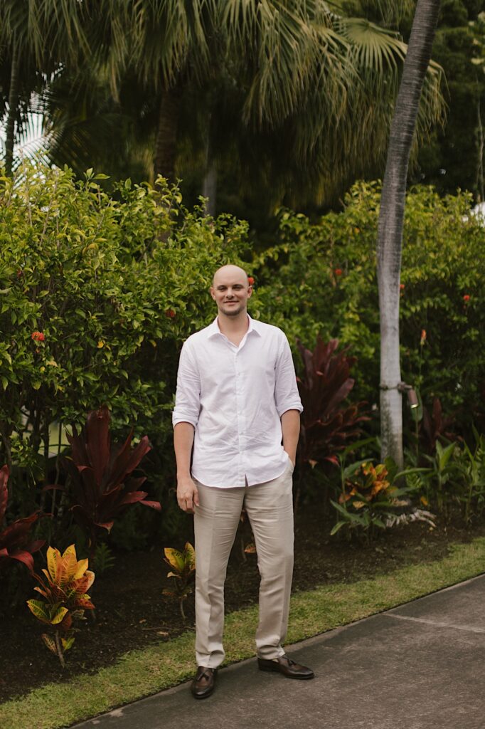 A groom stands and smiles at the camera in front of lush greenery of the Hawaiian islands