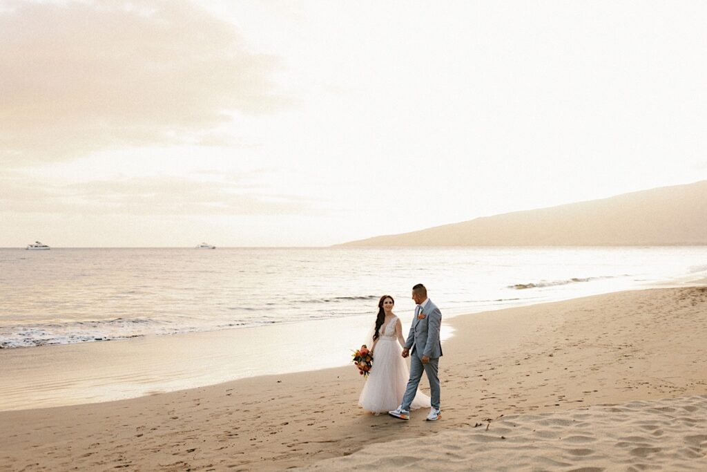 A bride and groom walk hand in hand while looking at one another along a beach on Maui during sunset