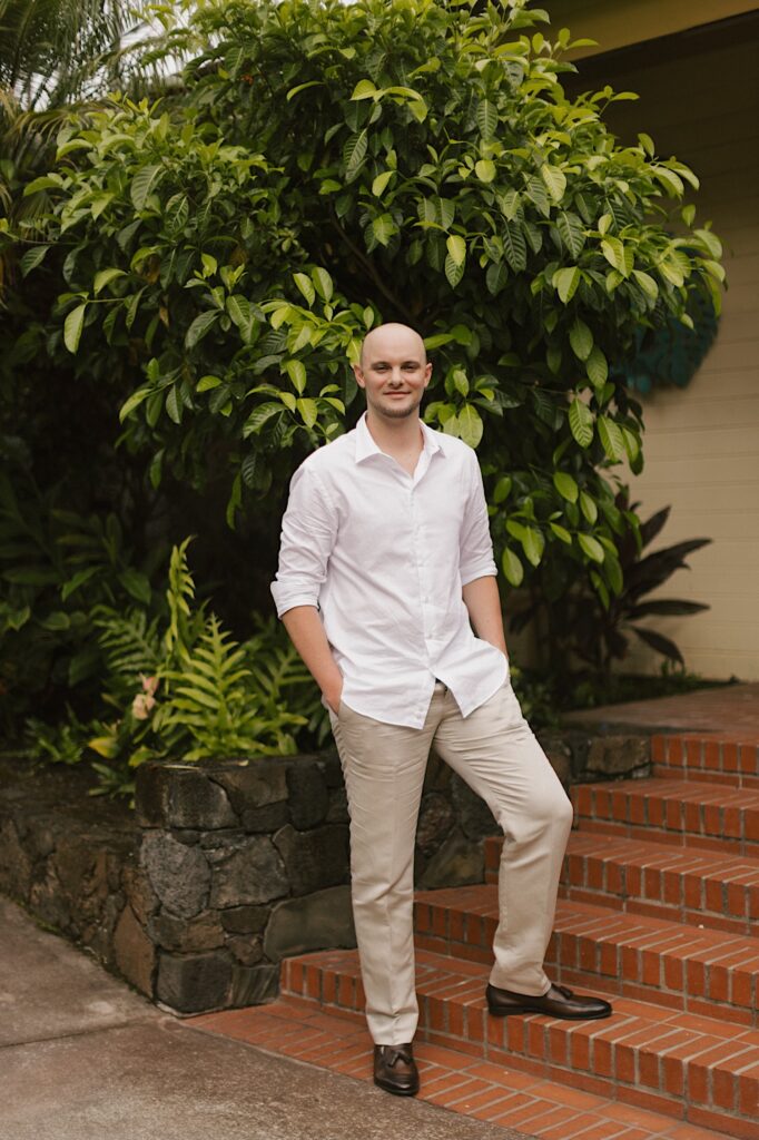 A groom smiles at the camera while standing on a brick staircase in front of a large green bush