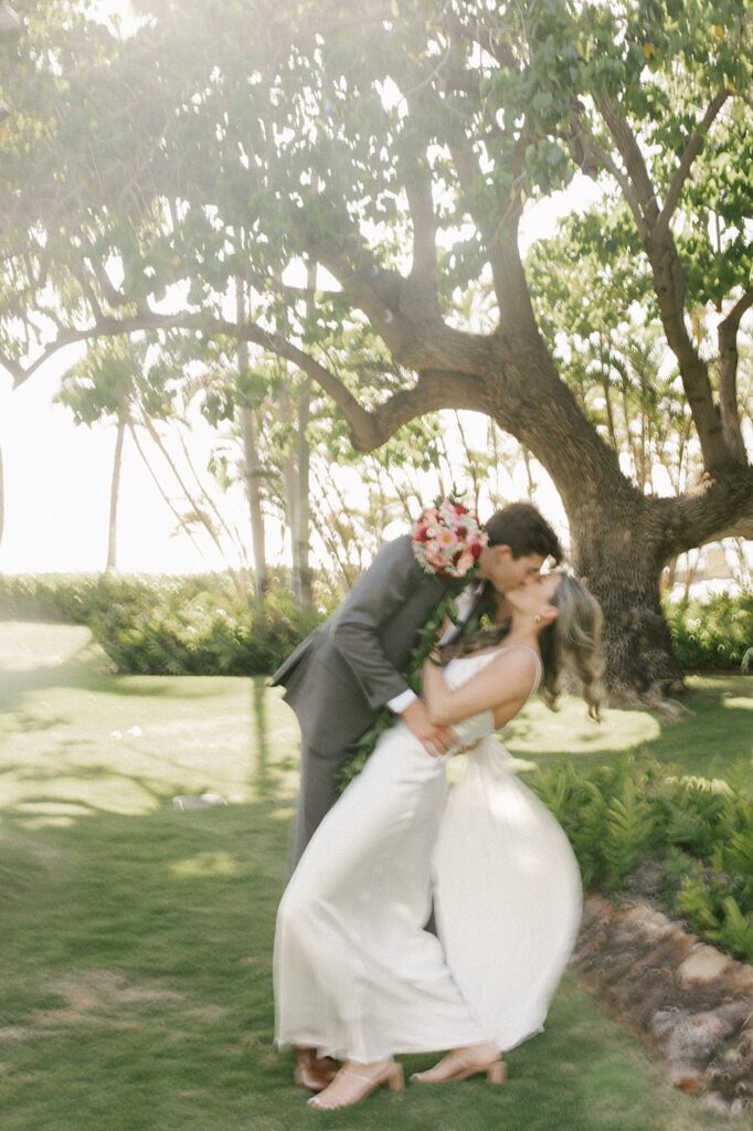 Blurry photo of a bride and groom kissing one another in front of a large tree, the bride has her arm wrapped around the groom's shoulder and is holding a bouquet