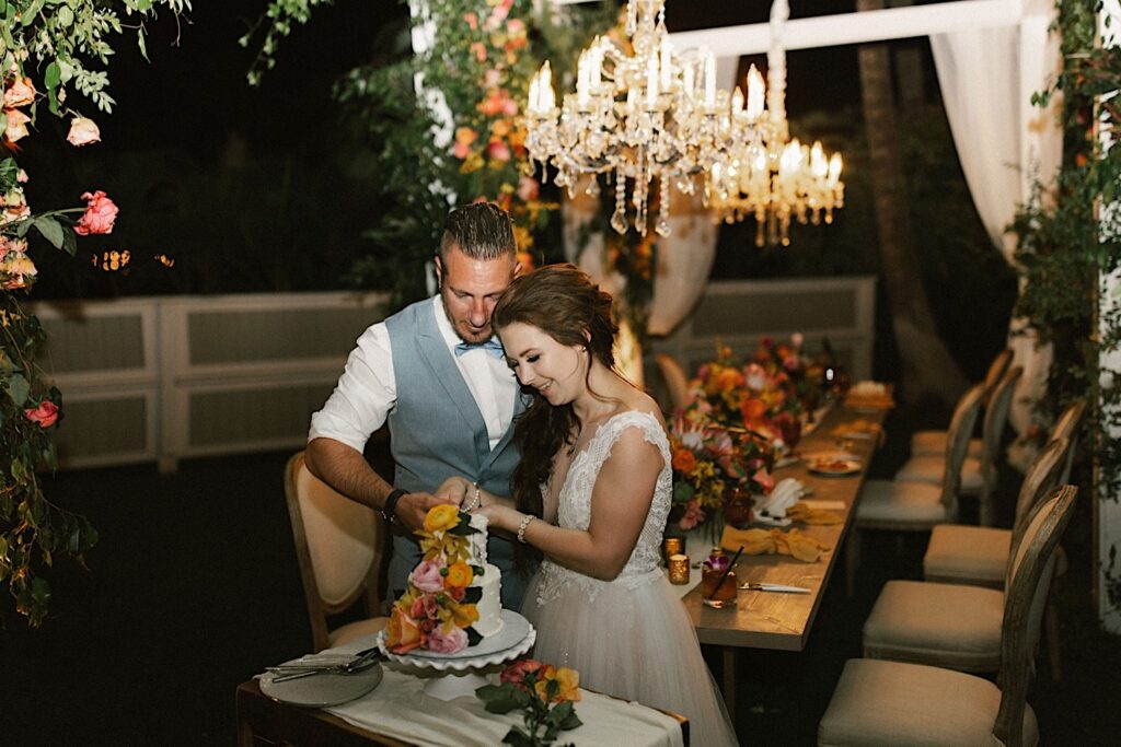 A bride and groom stand next to one another while cutting their wedding cake that is covered in flowers during their intimate wedding reception at their venue in Maui, behind them are chandeliers, a table and the rest of the outdoor reception space