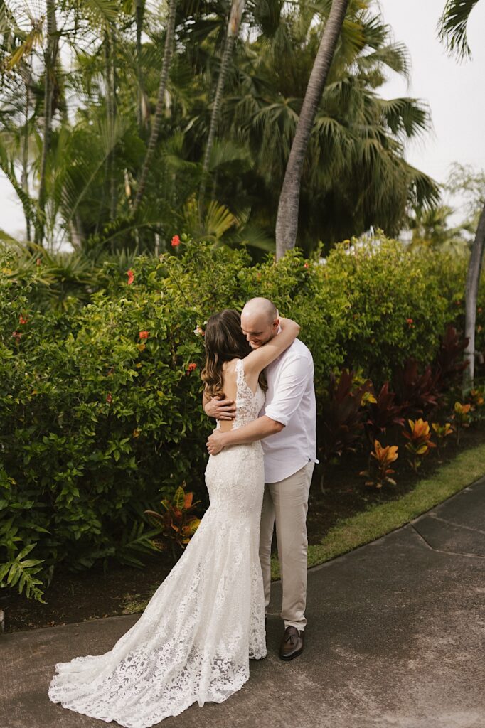 A bride and groom hug one another on a path in front of green bushes and palm trees after seeing each other for the first time