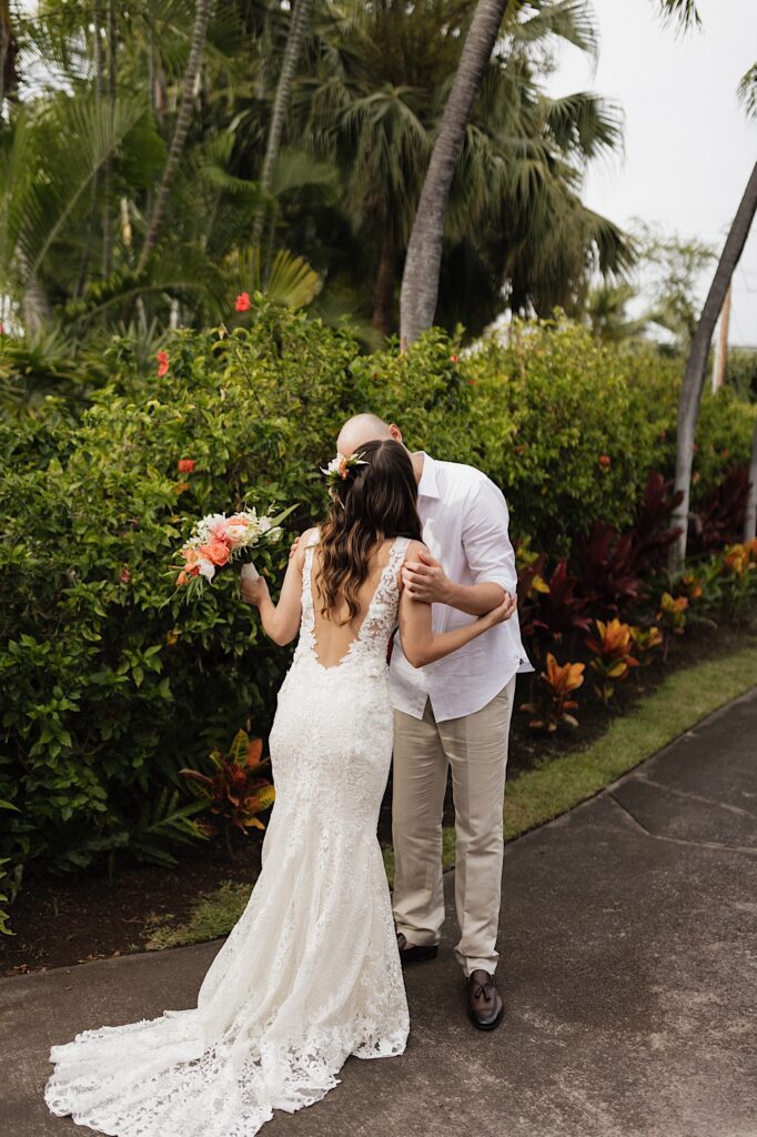 A bride and groom kiss one another on a path in front of green bushes and palm trees after seeing each other for the first time