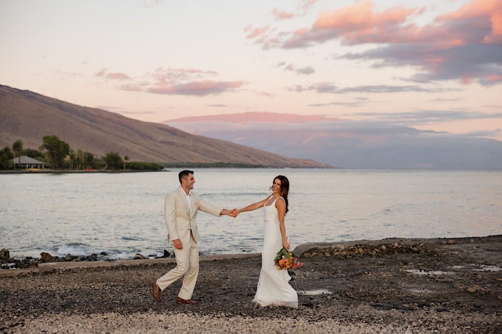 A bride and groom hold hands and smile while dancing on the beach looking out over the ocean and the Hawaiian islands during sunset