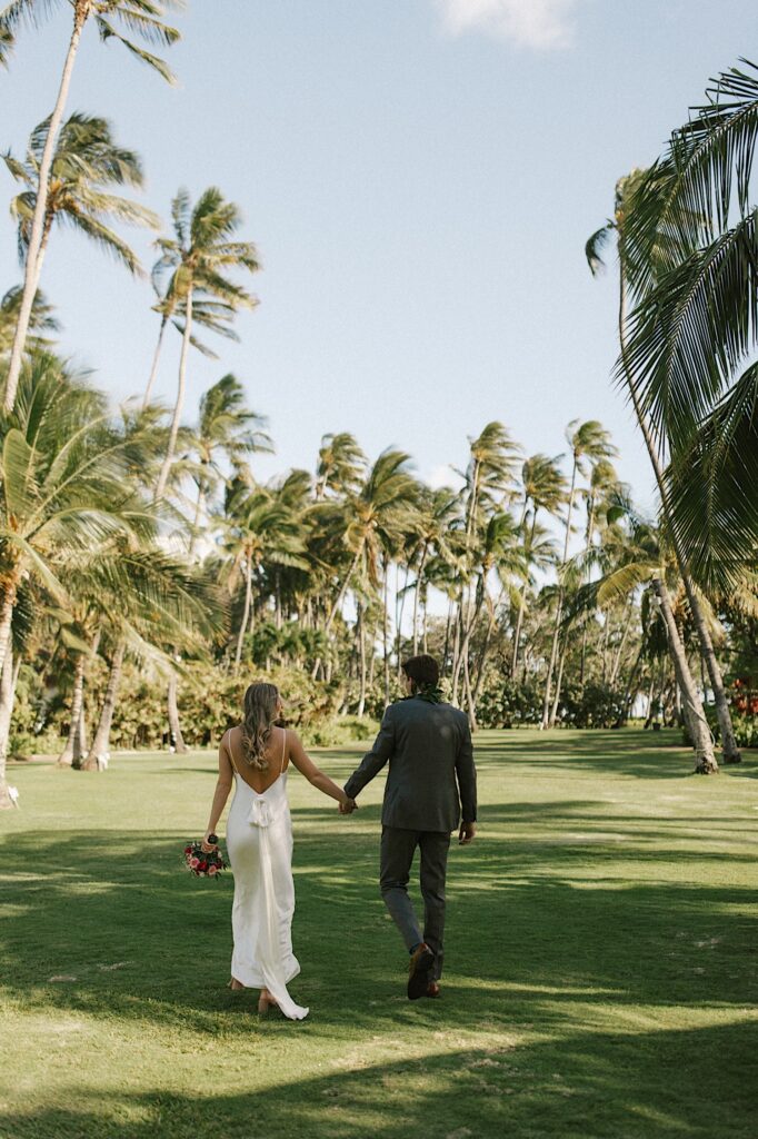 A bride and groom walk hand in hand away from the camera in a grass field towards palm trees
