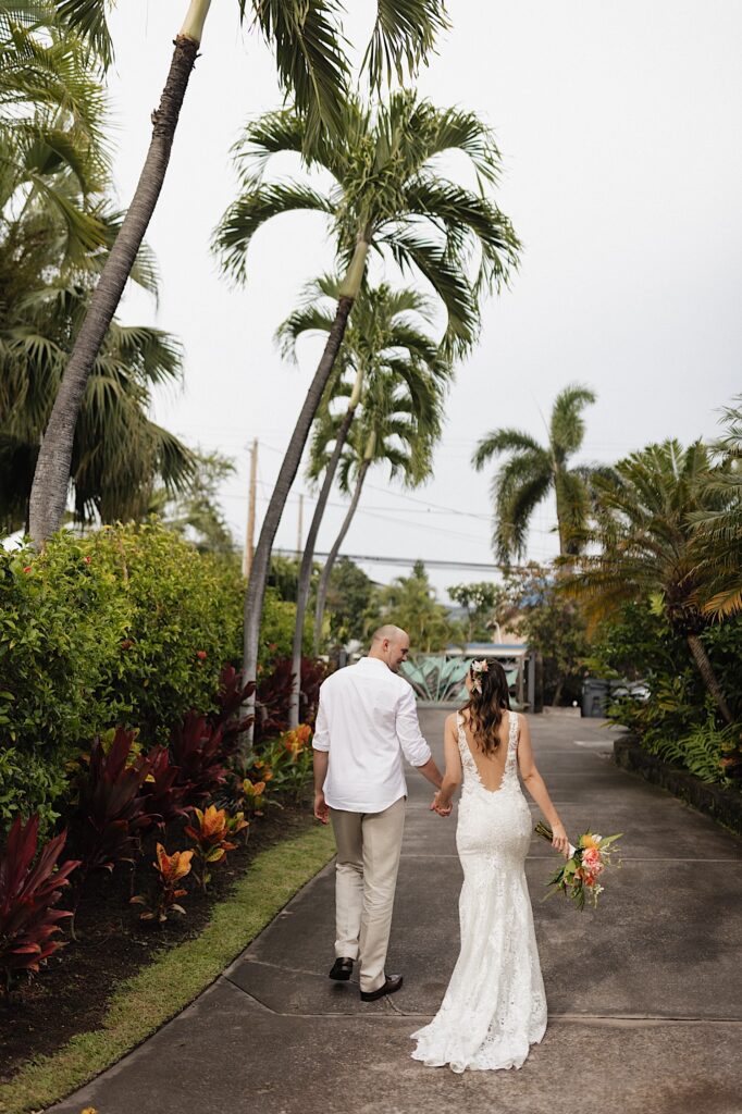 A bride and groom walk away from the camera hand in hand and look at one another along a path of palm trees and lush Hawaiian greenery