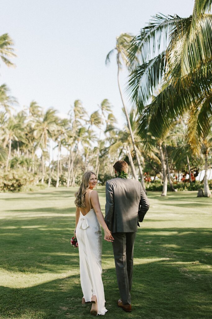 A bride and groom walk hand in hand away from the camera while the bride smiles over her shoulder to the camera, they are walking in a grass field towards palm trees