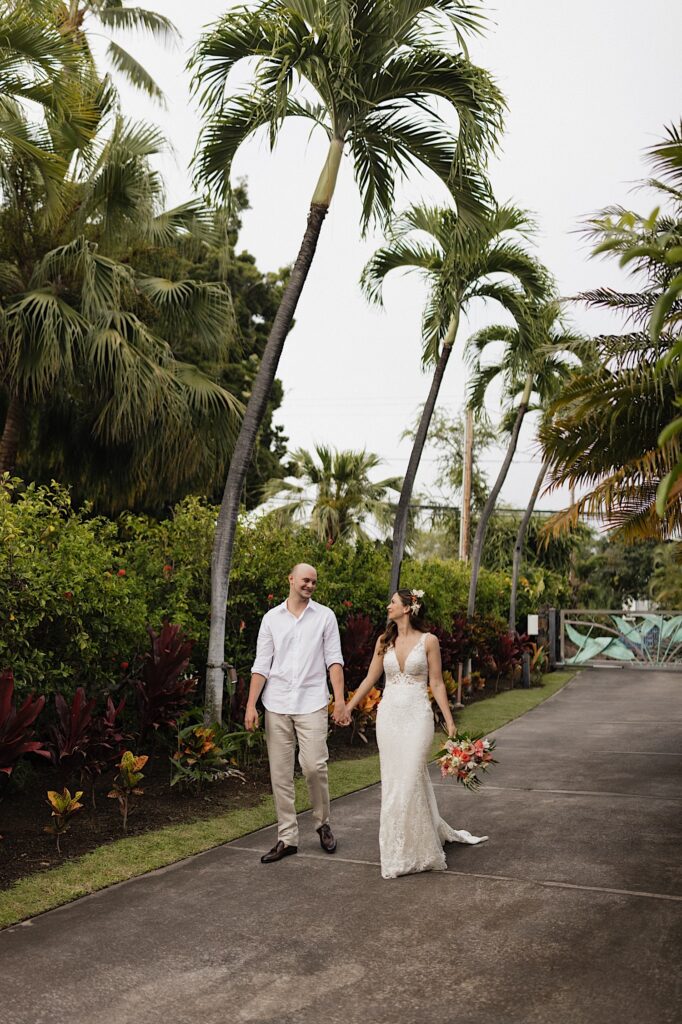 A bride and groom walk hand in hand towards the camera while smiling at one another along a path lined with lush greenery and palm trees
