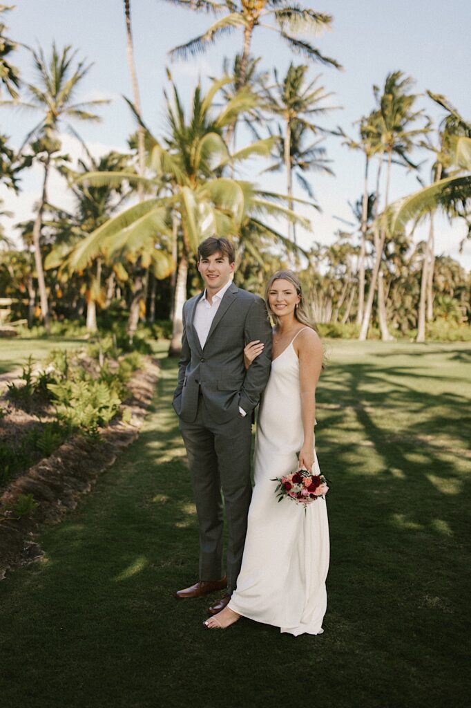 A bride and groom stand next to one another and smile at the camera while the bride holds the groom's arm, behind them are palm trees