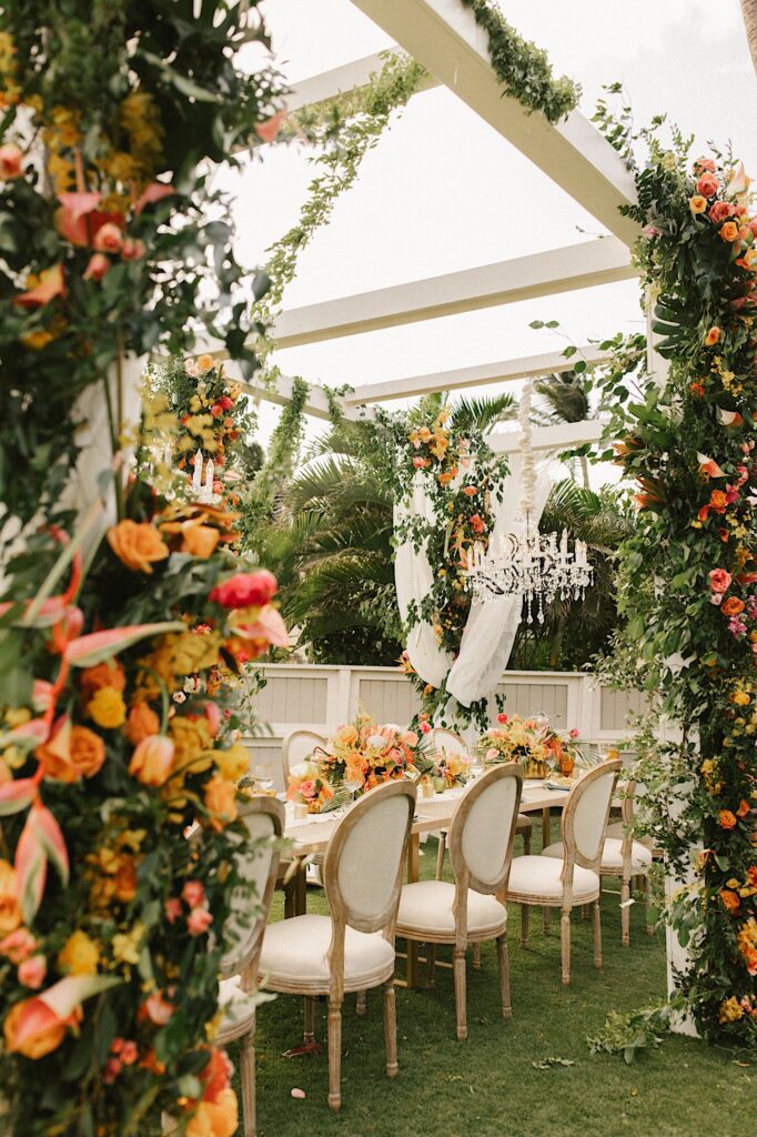 An outdoor wedding reception space decorated with tons of orange and pink flowers and a chandelier hanging from the outdoor overhang